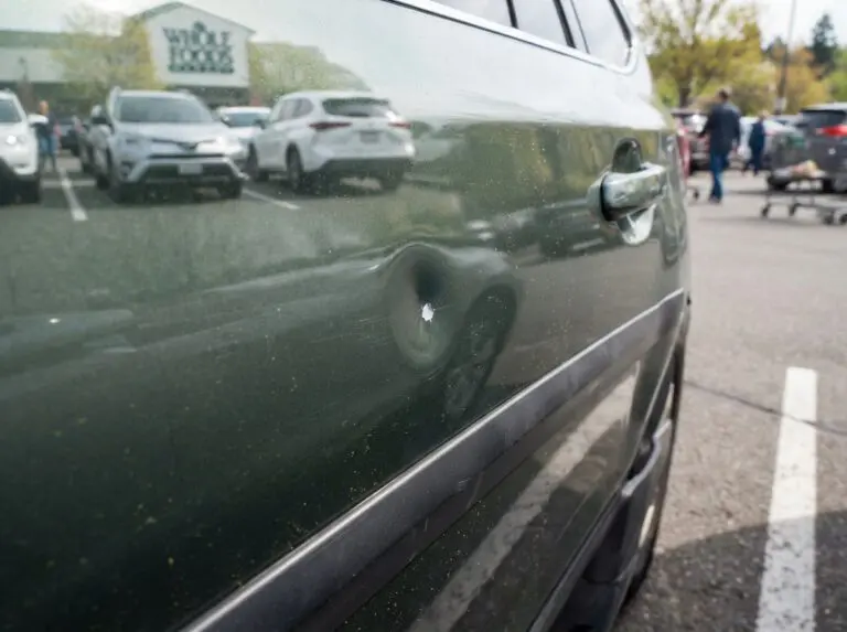 Kleine Delle mit Lackabplatzer an der Tür eines dunkelgrünen Autos auf einem Supermarktparkplatz – typischer Parkplatzschaden durch eine geöffnete Autotür.