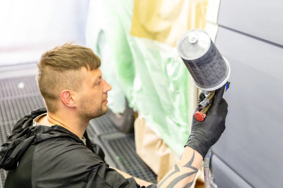 Fachkraft mit Tätowierung führt eine Autolackierung durch, hält eine Lackierpistole auf ein abgeklemmtes Fahrzeugteil in der Grünert Werkstatt.
